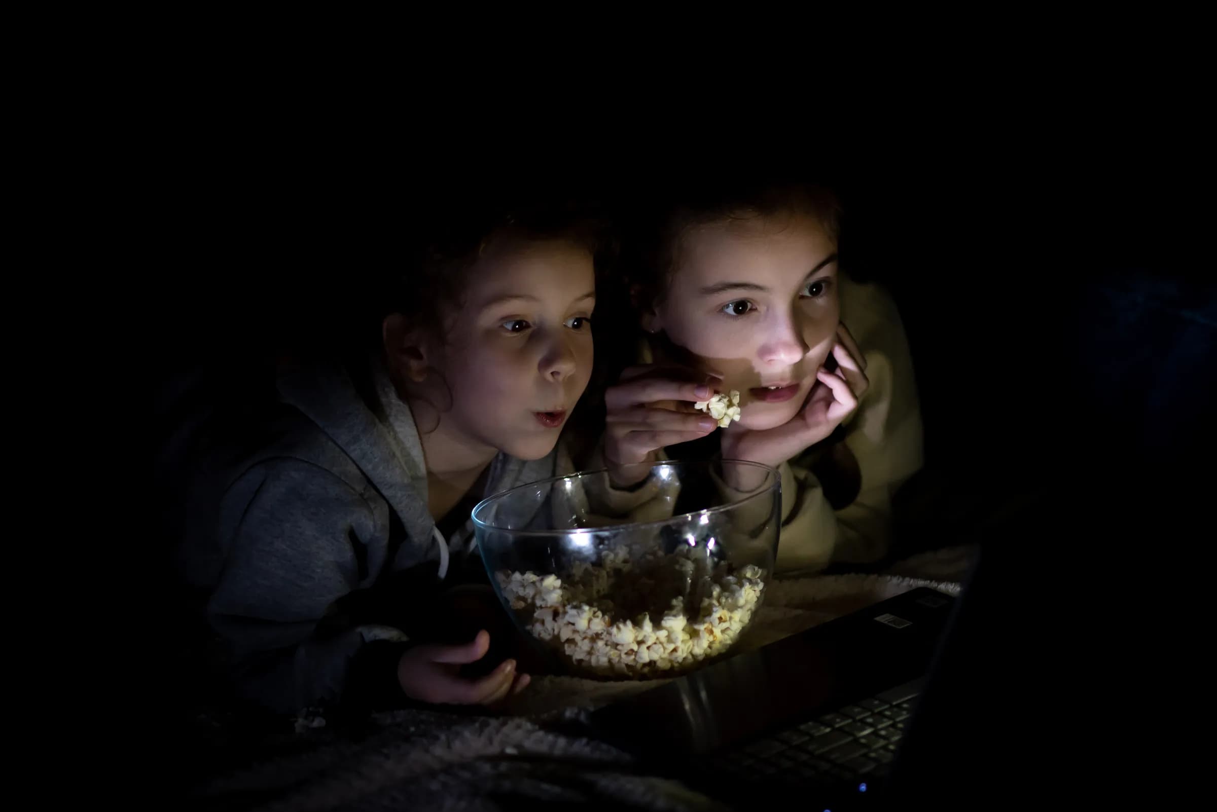 Two kids watching a movie at home at night, sharing popcorn